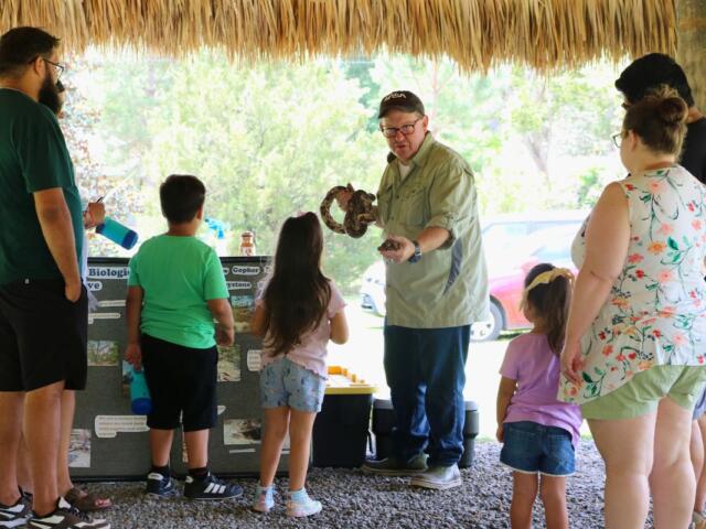 A man demonstrates a tortoise and snake to children and families under a chickee.