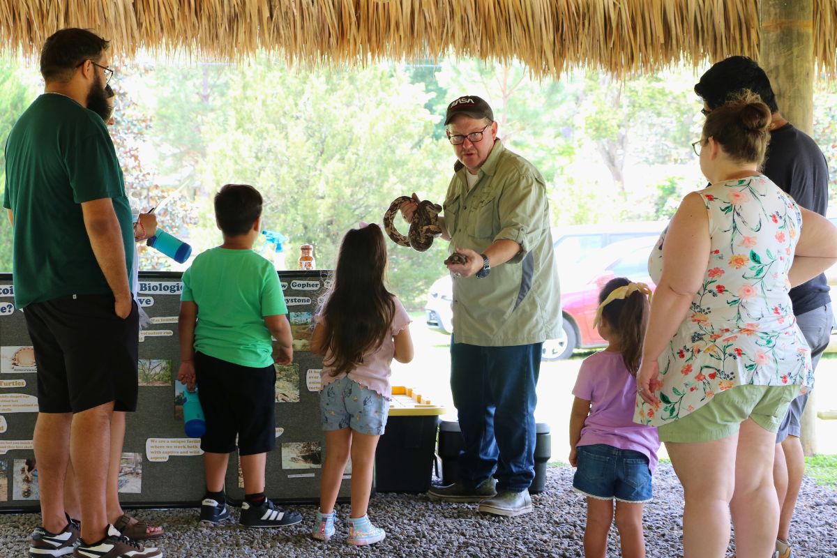A man demonstrates a tortoise and snake to children and families under a chickee.