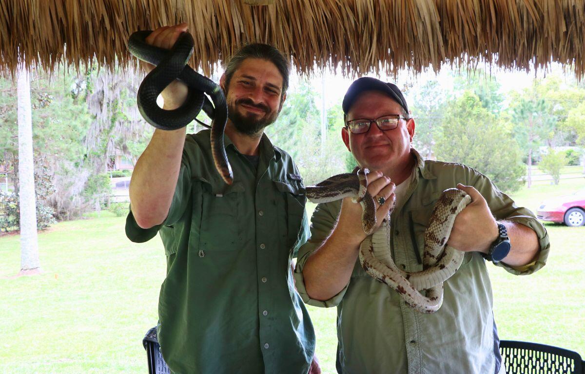 Two men holds snakes in their hands in a wooded area under a chikee.