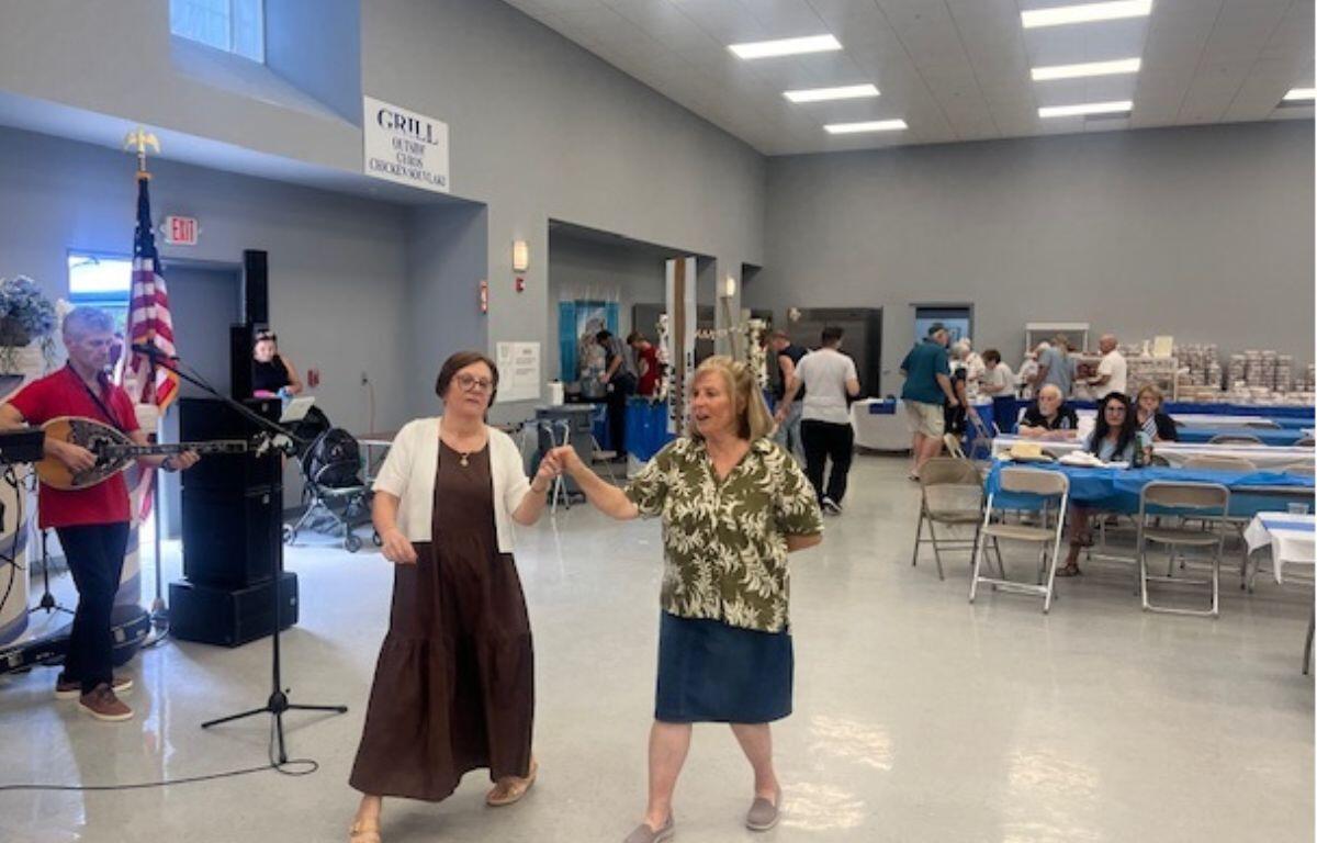 On the left a man playing a bouzouki and a speaker, with two women as the featured image dancing, with people sitting at tables in the background.