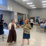 On the left a man playing a bouzouki and a speaker, with two women as the featured image dancing, with people sitting at tables in the background.