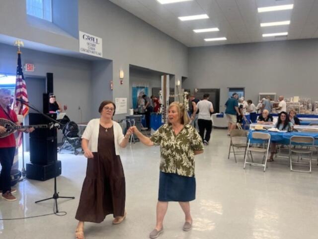 On the left a man playing a bouzouki and a speaker, with two women as the featured image dancing, with people sitting at tables in the background.