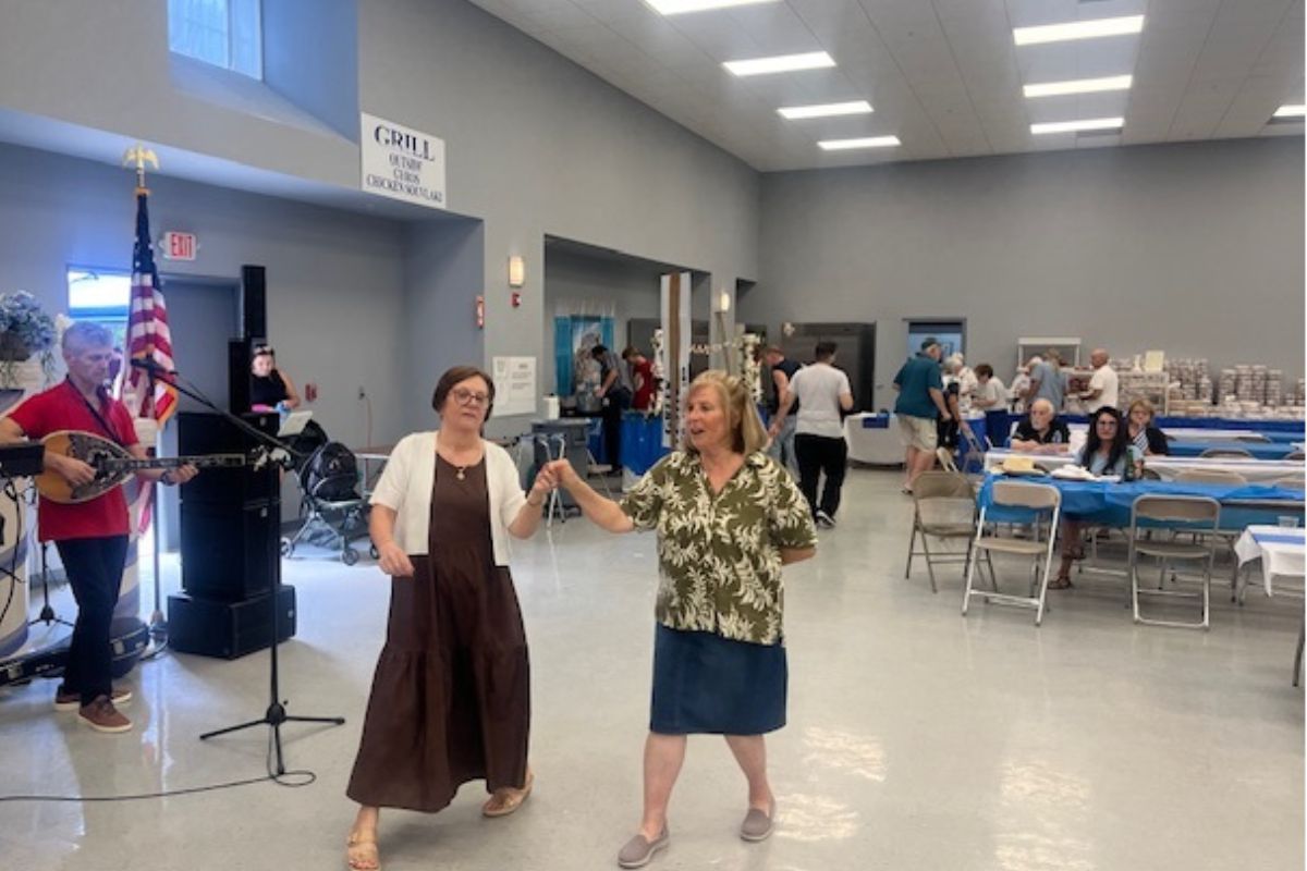 On the left a man playing a bouzouki and a speaker, with two women as the featured image dancing, with people sitting at tables in the background.
