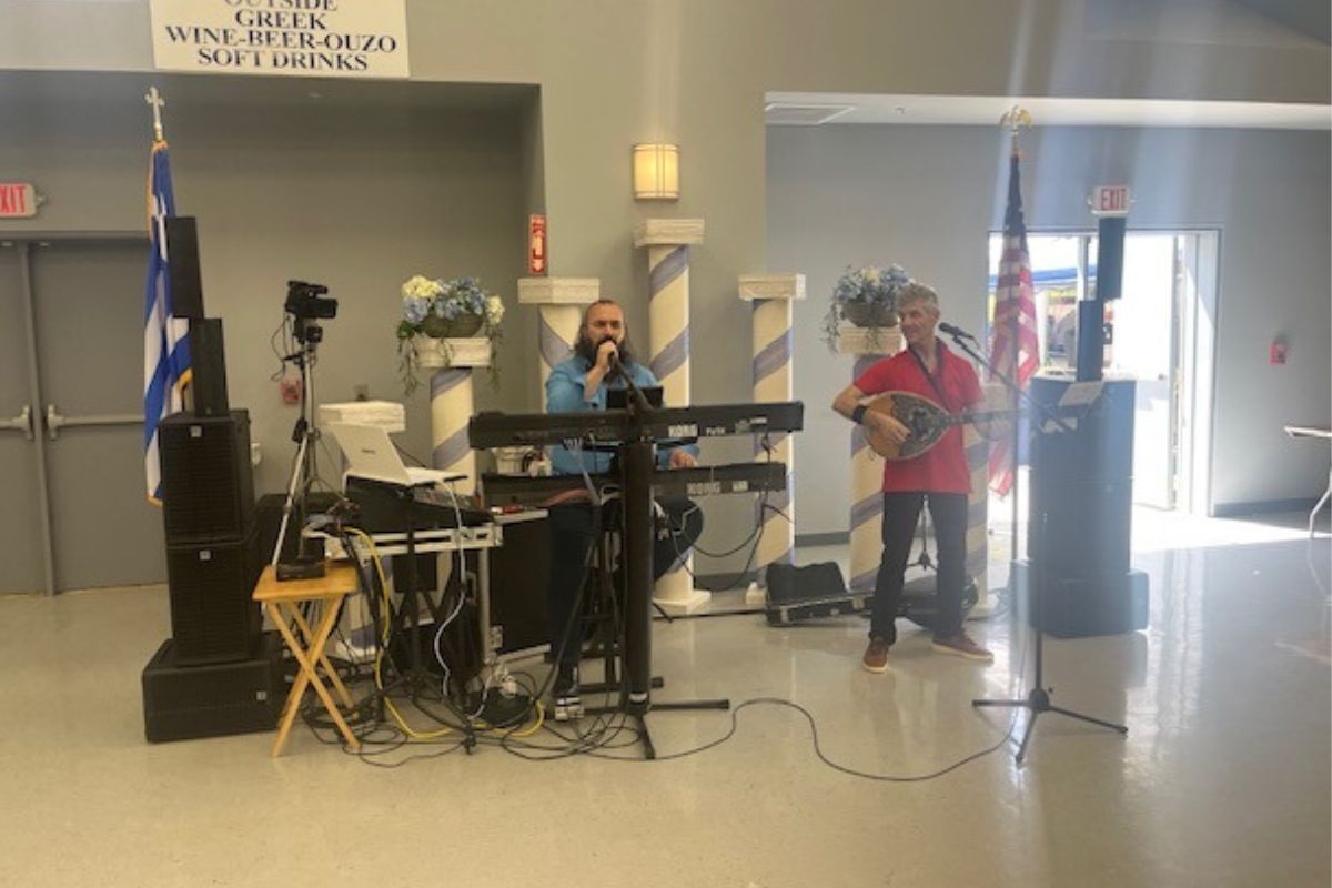 A Greek Flag on the extreme left, speaker, a keyboard, a man behind the keyboard singing, to the right, a man playing a bouzouki and the flag of the United States of America. 