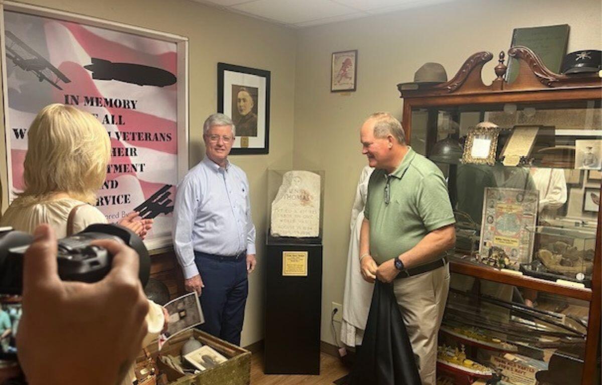 People standing to the left in the foreground, A frame of an American Flag poster to the left, two men standing on either side of a headstone on a pedestal, with the man on the left standing in front of framed picture and the man on the right standing in front of secretary with a slew of items.