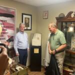People standing to the left in the foreground, A frame of an American Flag poster to the left, two men standing on either side of a headstone on a pedestal, with the man on the left standing in front of framed picture and the man on the right standing in front of secretary with a slew of items.