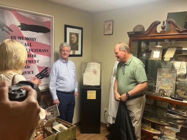 People standing to the left in the foreground, A frame of an American Flag poster to the left, two men standing on either side of a headstone on a pedestal, with the man on the left standing in front of framed picture and the man on the right standing in front of secretary with a slew of items.