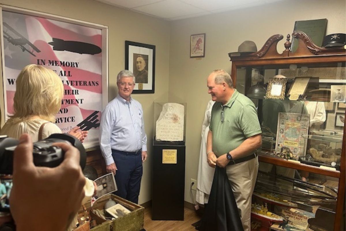 People standing to the left in the foreground, A frame of an American Flag poster to the left, two men standing on either side of a headstone on a pedestal, with the man on the left standing in front of framed picture and the man on the right standing in front of secretary with a slew of items.