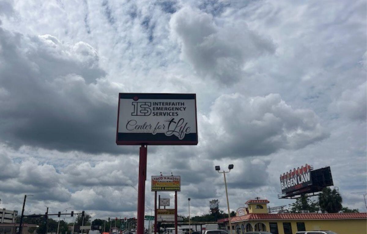 To the left cars and a traffic signal, blue skies and clouds over head, a building with cars in a parking lot to the right, extreme right a billboard. An elevated sign in the foreground.