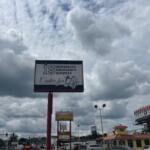 To the left cars and a traffic signal, blue skies and clouds over head, a building with cars in a parking lot to the right, extreme right a billboard. An elevated sign in the foreground.