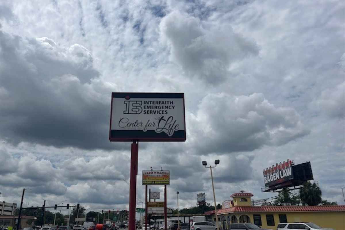 To the left cars and a traffic signal, blue skies and clouds over head, a building with cars in a parking lot to the right, extreme right a billboard. An elevated sign in the foreground.