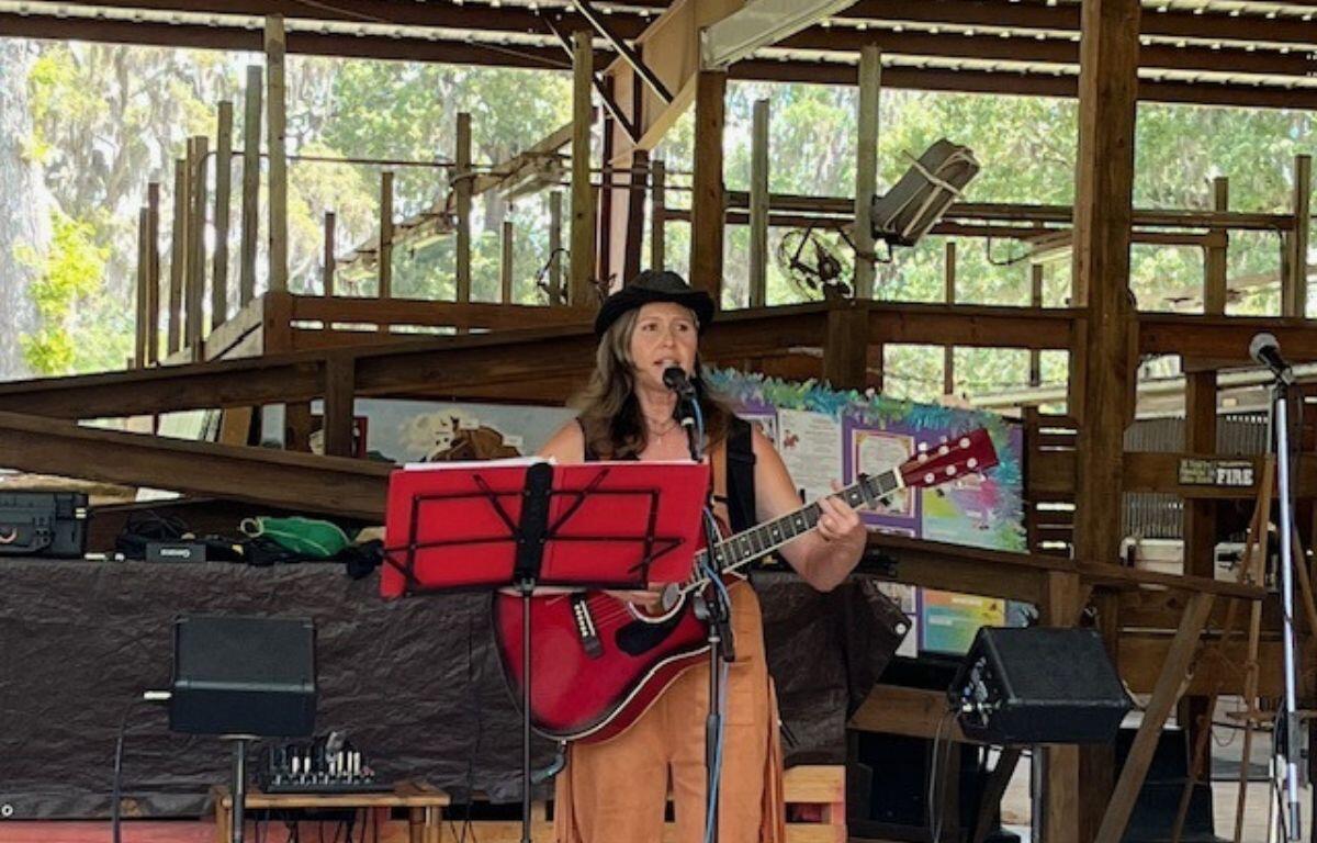 A woman wearing a hat playing the guitar and singing in an open-aired barn, standing in front of a music stand, with a speaker to her right.