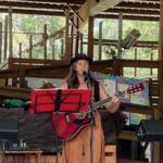 A woman wearing a hat playing the guitar and singing in an open-aired barn, standing in front of a music stand, with a speaker to her right.