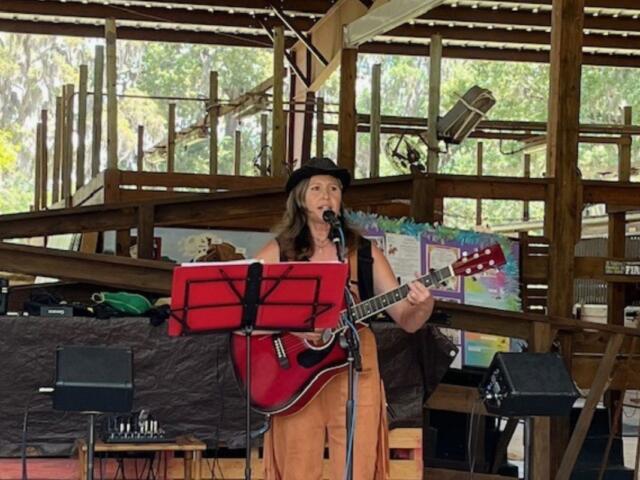 A woman wearing a hat playing the guitar and singing in an open-aired barn, standing in front of a music stand, with a speaker to her right.