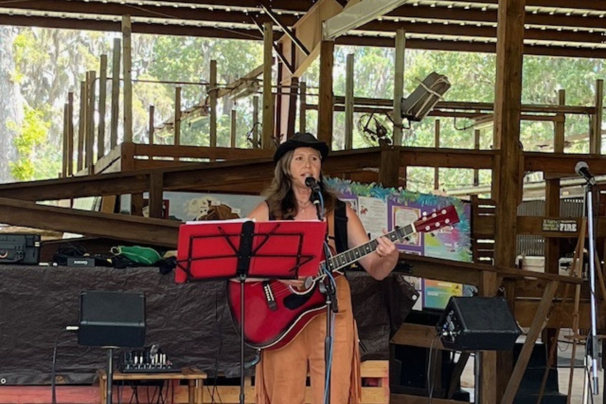 A woman wearing a hat playing the guitar and singing in an open-aired barn, standing in front of a music stand, with a speaker to her right.
