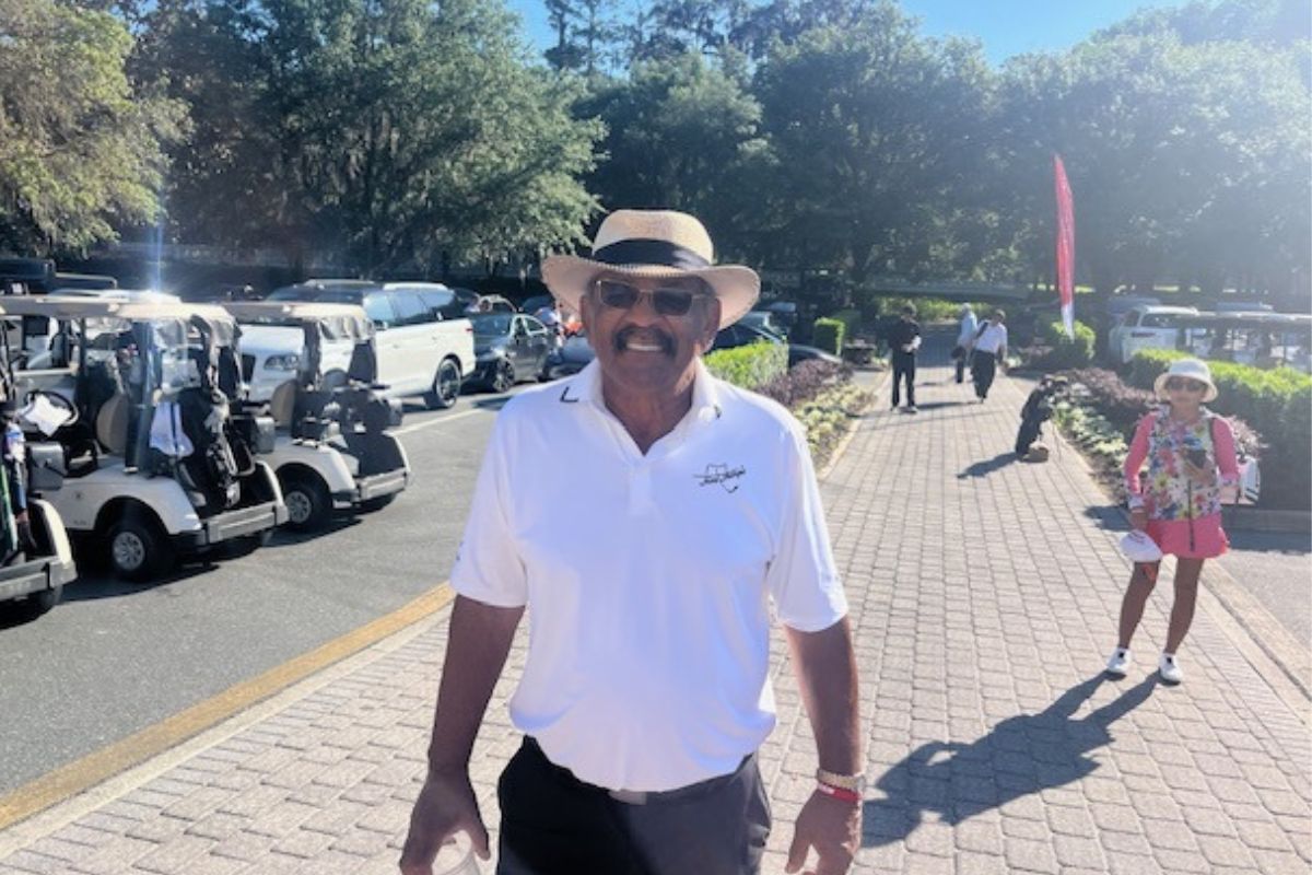 Golf carts and vehicles to the left, in the background on a brick road, people walking, and the featured image, a man wearing a hat with sunglasses.