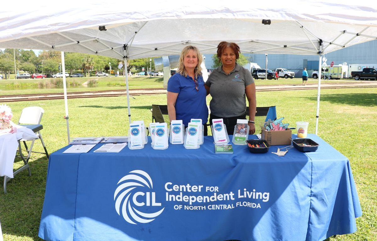 Two women stand behind a table in a park with a lake in the background.