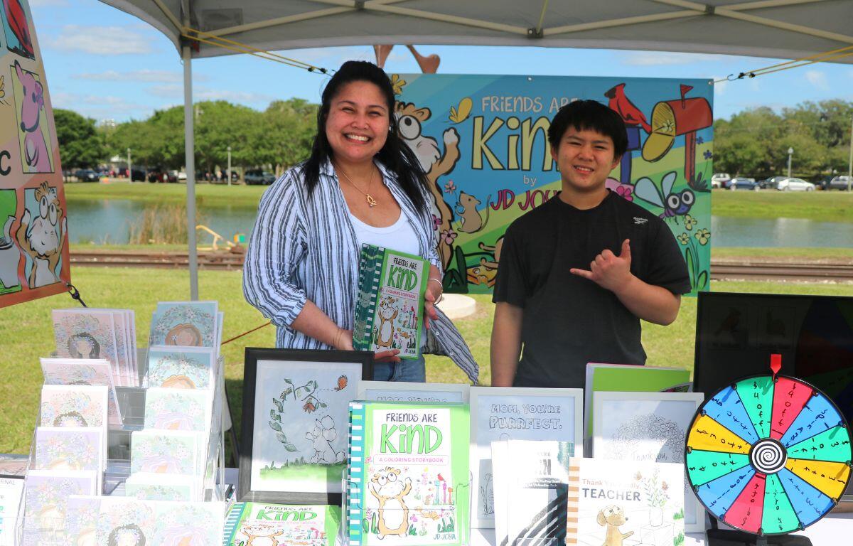 A mother and son stand behind a table filled with books and cards. Behind them is a banner that reads, "Friends are Kind by JD."