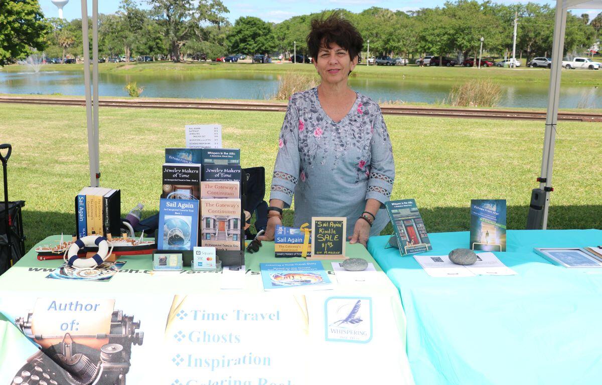 A woman stands behind a table with books scattered around. Behind her are railroad tracks and a lake.