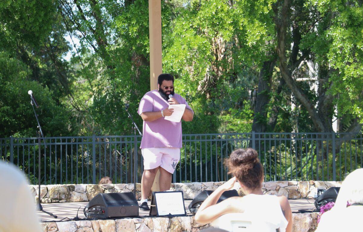 A man stands on a stage with a piece of paper, reading his poetry to an audience of people.