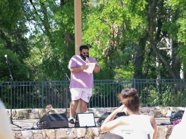 A man stands on a stage with a piece of paper, reading his poetry to an audience of people.