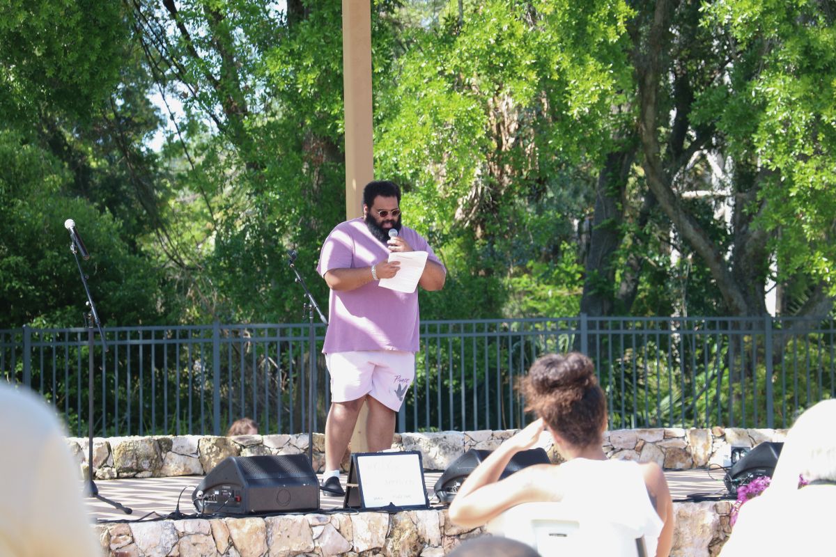 A man stands on a stage with a piece of paper, reading his poetry to an audience of people.