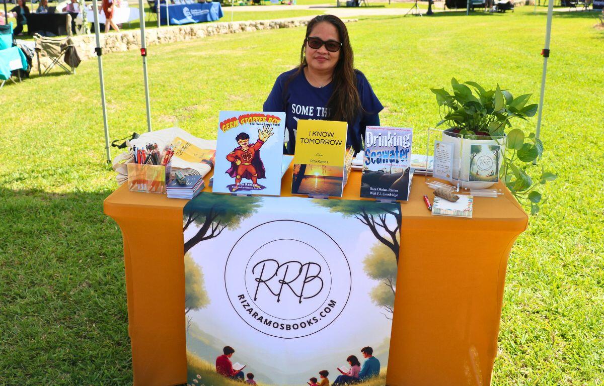 A women sits at a small stand with books on top of it. Behind her is a literary festival and market.
