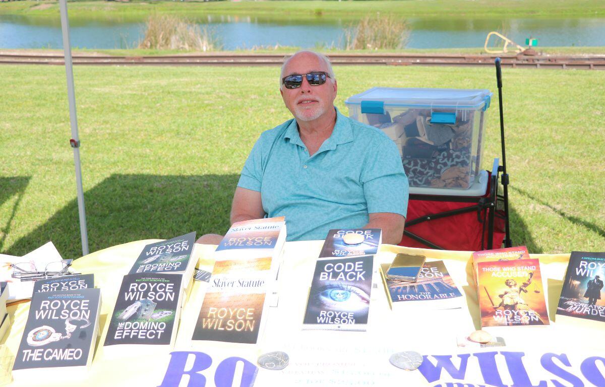 A man sits behind a table with books scattered around. Behind him is train tracks and a lake.