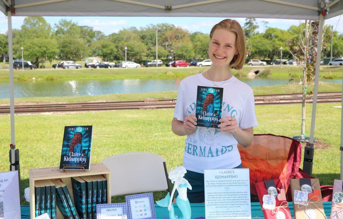 A girl holds her book behind a table in front of a lake.