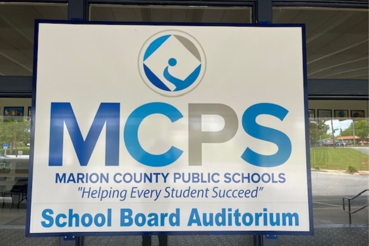 A sign attached to a window reflecting the parking lot. The sign itself has writing featuring different sizes, fonts and colors.