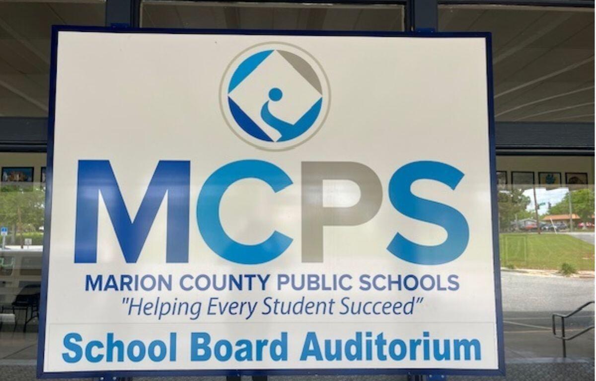 A sign with lettering of various sizes and a logo, against a series of windows, with the reflection of images within the parking lot.