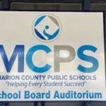 A sign with lettering of various sizes and a logo, against a series of windows, with the reflection of images within the parking lot.