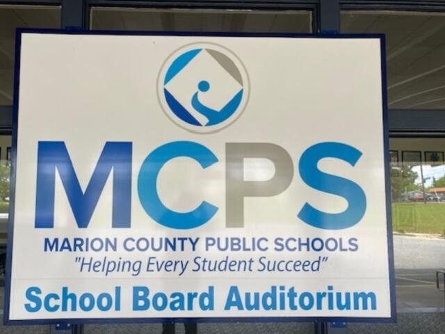 A sign with lettering of various sizes and a logo, against a series of windows, with the reflection of images within the parking lot.