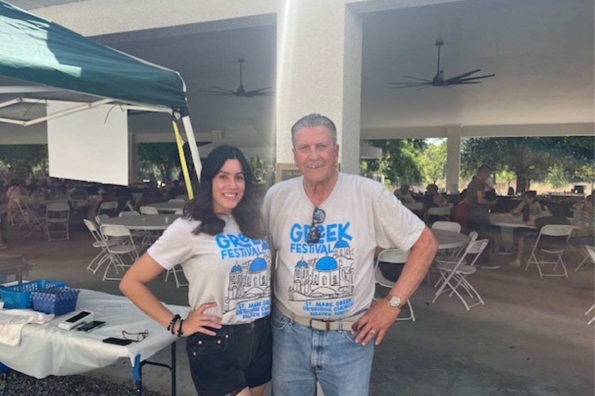 A tent with a table under it, a woman an man standing in front of a column, with people in the background at tables. 