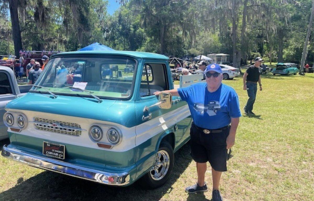 On the left two vehicles, a man to the right of the vehicle. In the background live oak trees with Spanish moss hanging off of them, a tent in the background and a man walking to the right on a grass field.