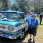 On the left two vehicles, a man to the right of the vehicle. In the background live oak trees with Spanish moss hanging off of them, a tent in the background and a man walking to the right on a grass field.