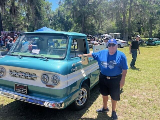 On the left two vehicles, a man to the right of the vehicle. In the background live oak trees with Spanish moss hanging off of them, a tent in the background and a man walking to the right on a grass field.