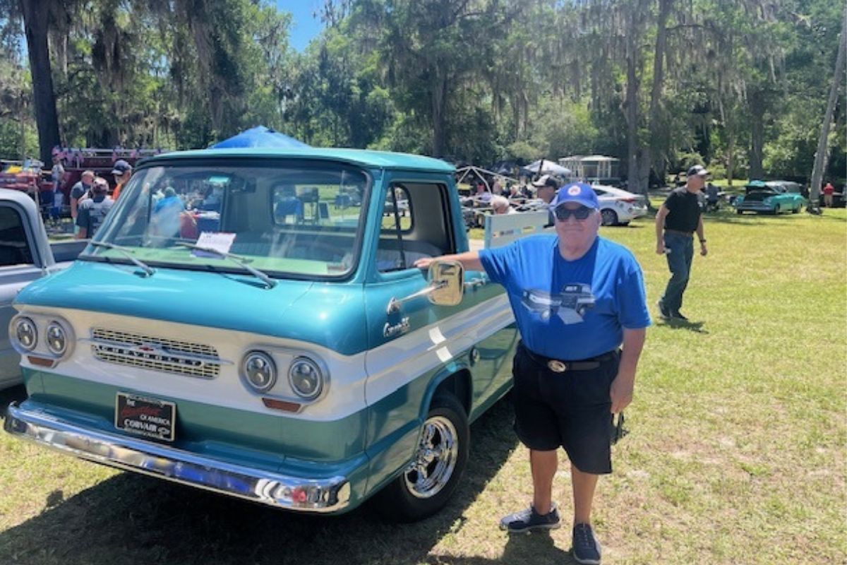 On the left two vehicles, a man to the right of the vehicle. In the background live oak trees with Spanish moss hanging off of them, a tent in the background and a man walking to the right on a grass field.