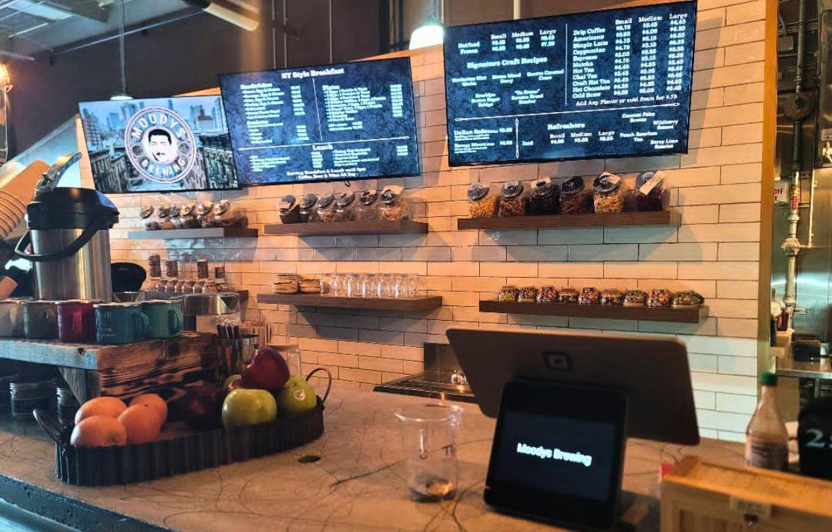 A coffee shop features a small kiosk on a counter with fruits next to it in a tray. Behind the counter are coffee items with large screens above it reading the menu.