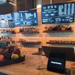 A coffee shop features a small kiosk on a counter with fruits next to it in a tray. Behind the counter are coffee items with large screens above it reading the menu.