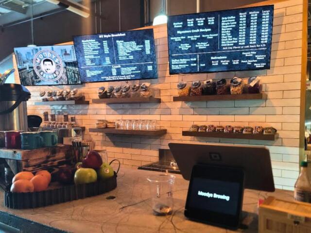 A coffee shop features a small kiosk on a counter with fruits next to it in a tray. Behind the counter are coffee items with large screens above it reading the menu.