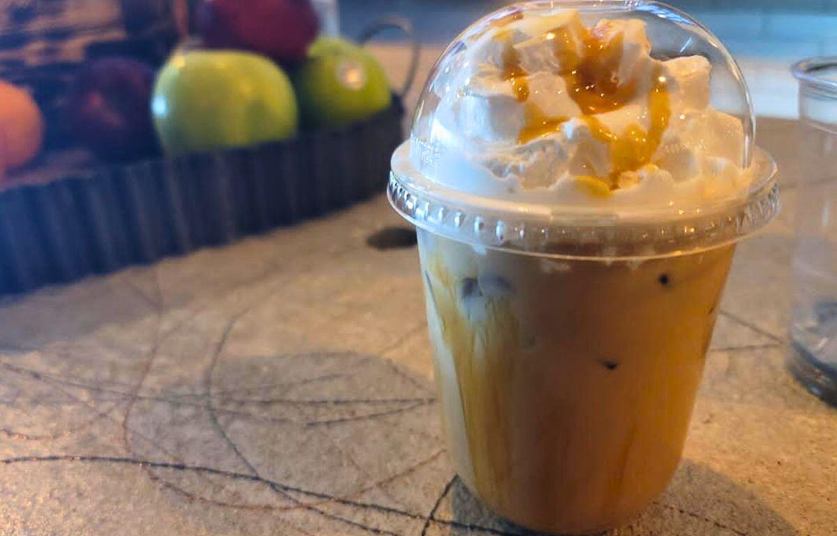 A coffee in a plastic container sits on a cashier counter. A carrier with apples sits in the background.