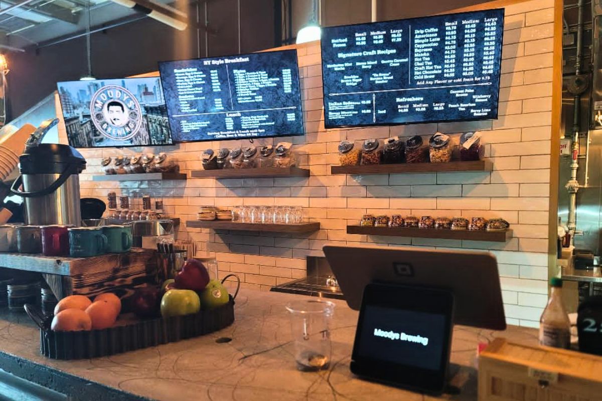 A coffee shop features a small kiosk on a counter with fruits next to it in a tray. Behind the counter are coffee items with large screens above it reading the menu.