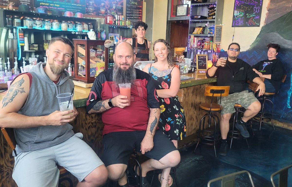 Six friends pose at a colorful bar, holding drinks and smiling at the camera with shelves and a chalkboard menu in the background.