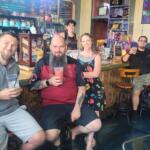 Six friends pose at a colorful bar, holding drinks and smiling at the camera with shelves and a chalkboard menu in the background.