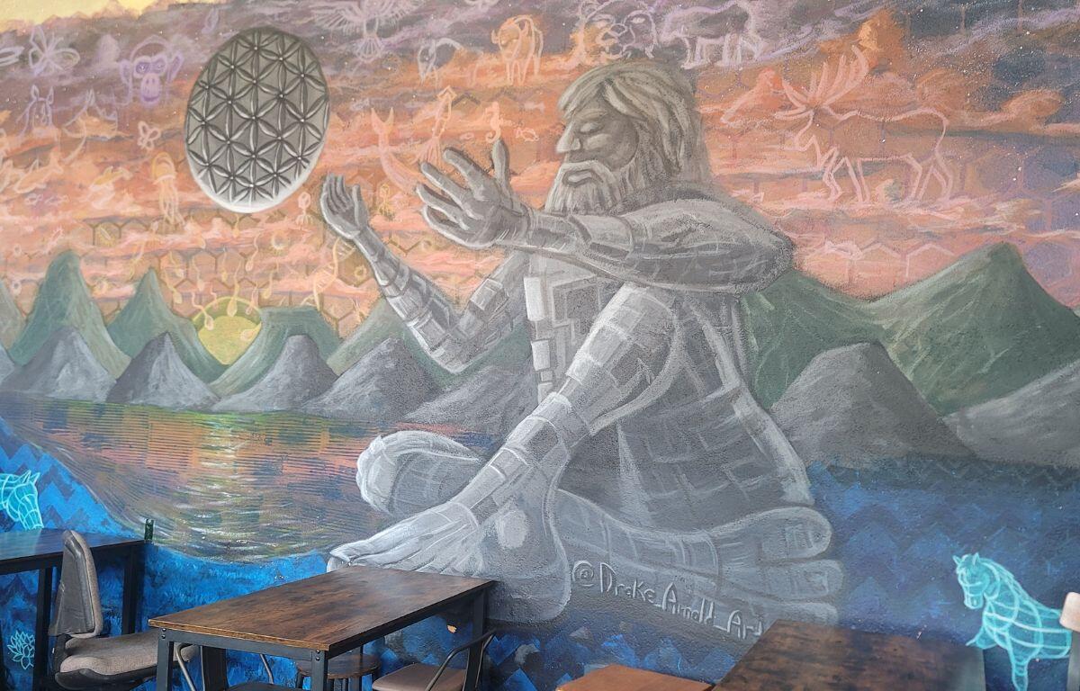 Indoor room with a large wall mural of a stone-like figure sitting cross-legged, meditating, reaching toward a geometric floating ball over a mountainous lake scene; foreground tables and chairs.