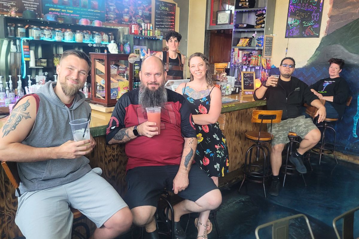 Six friends pose at a colorful bar, holding drinks and smiling at the camera with shelves and a chalkboard menu in the background.