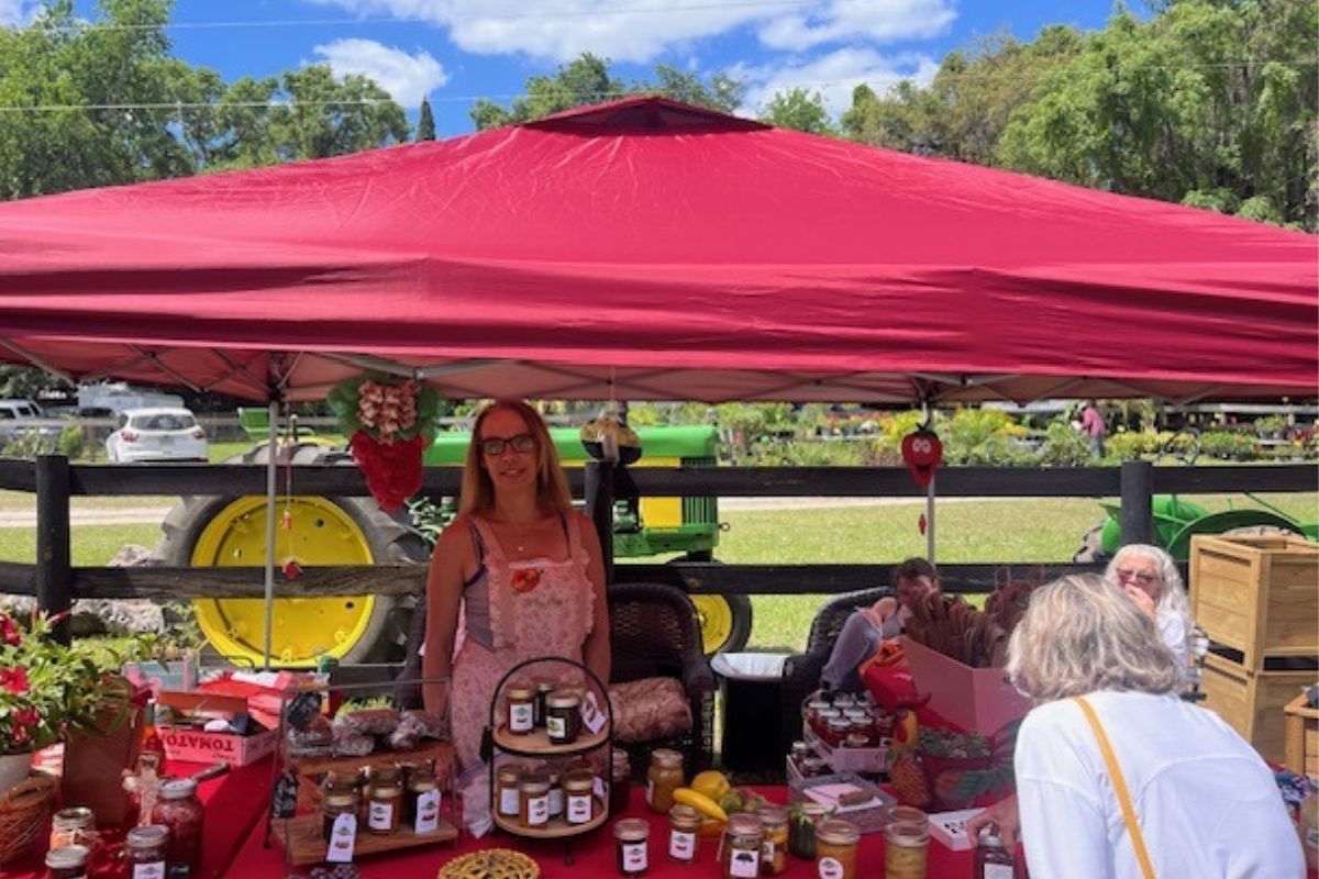 A woman in the center of the photo, standing under a red tent canopy, with paddock fencing in the background, a tractor, green space and a tree line. In the foreground mason jars and a woman leaning over to the right.
