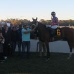 People to the left, with a racetrack, people and a tree line in the background, with a horse and rider up to the right.