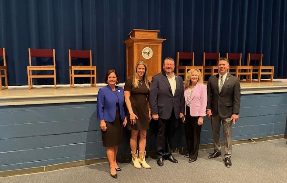 A gray concrete block base to a stage in the background, with chairs to the right and left of a podium. Well-dressed people standing together in front of the stage.